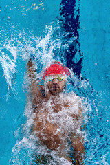 Fototapeta premium Top view of professional male swimmer in red swimming cap and goggles practicing and training at pool, indoors. Healthy lifestyle, power, energy, sports movement concept
