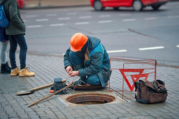 Utility worker sits in front of an open sewer hatch on city street. Electricity and communications...