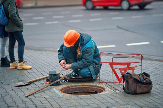 Utility worker sits in front of an open sewer hatch on city street. Electricity and communications repair, underground utilities, cable laying works.