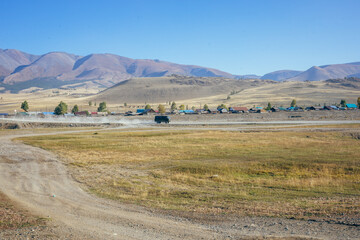 UAZ goes through the rural steppe. SUV in the village. Off-road, dust from a passing car. Village houses. Mountains, blue sky.