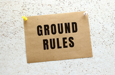 A sheet of craft paper with GROUND RULES text attached to a white textured wall with a button. Office reminder.