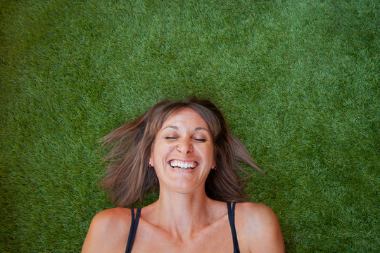 Mujer sonriendo echada sobre su espalda en el cesped verde. Mujer de pelo marr&oacute;n y camiseta de tirantes negro. Retrato de chica