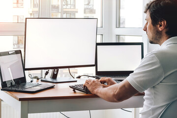 Man sitting at workplace with two laptops and monitor near the window. Remote work from home