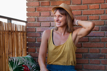 Mujer sentada en la terraza de su casa en un sofá hecho con palets, con cojines de flores y plantas, con valla de bambú, suelo de cesped y de fondo una pared de ladrillos. Chica sonriente y pensante