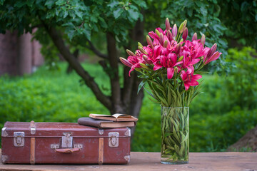 Beautiful bouquet of red lily flowers in a glass vase with water and old suitcase, book on a wooden table in garden