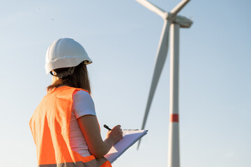 Woman in the orange vest makes notes on maintenance of wind turbines on the background of windmill...