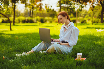 Beautiful young pretty redhead woman in park outdoors using laptop computer for study or work online, wireless earphones on her neck