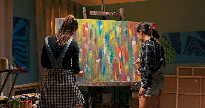Talented girls spend their free evening in an art studio, standing in front of a huge easel with a canvas on which they finish painting their colorful masterpiece, the last brush strokes