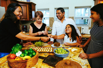 Cheerful aunt laughing and giving cheese to niece while enjoying with family in kitchen at home during weekend