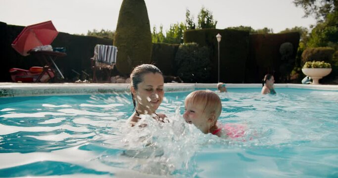 Young beautiful woman teaches her baby daughter to swim in swimming pool her son plays with his aunt at background. Family spending time together at summer vacations in rural house in Spain, Catalunya