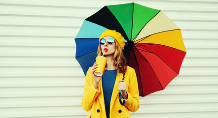 Autumn portrait of beautiful young woman with colorful umbrella and cup of juice wearing a yellow coat and beret on a white background