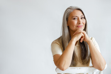 Happy middle aged Asian woman with loose silver hair sits on chair leaning on hands on light...