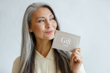 Positive silver haired Asian woman customer holds gift card standing on light grey background in...