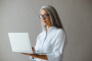 Concentrated long haired mature Asian woman wearing white blouse works on contemporary laptop...