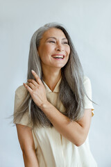 Positive middle aged Asian woman touches natural hoary hair poses for camera on light grey...
