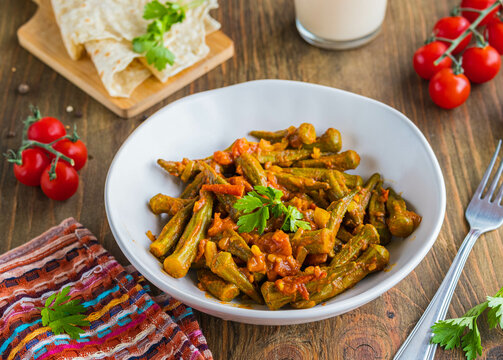 Stewed okra with tomatoes and spices in a clay bowl on a brown wooden background. Indian food.