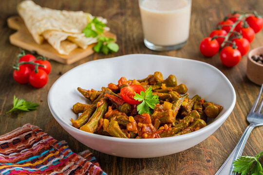 Stewed okra with tomatoes and spices in a clay bowl on a brown wooden background. Indian food.