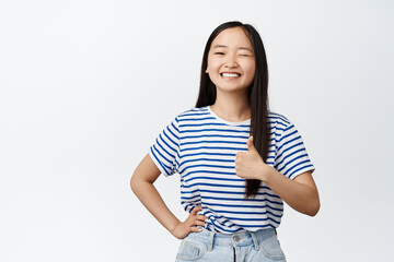 Smiling asian woman winking at camera, showing thumb up in approval, encourage you, praise good...