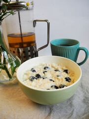 in a green round plate with oatmeal, coconut milk and blueberries, it lies on the table next to a teapot of green tea and a turquoise mug . side view