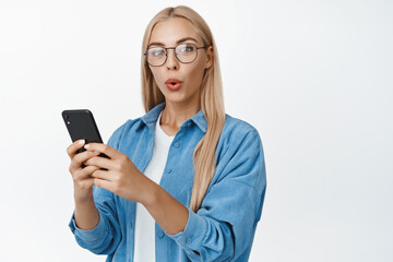 Fototapeta premium Portrait of surprised blond girl in glasses, holding smartphone and looking intrigued at camera, standing over white background
