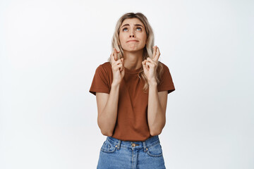 Nervous young woman praying, holding fingers crossed and making wish, standing over white background, hoping for dream come true