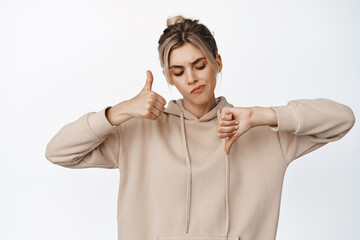 Image of doubtful blond girl showing thumbs up and down, looking at dislike gesture with thoughtful...