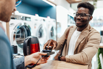 African American man buying electric water kettle in electronics and appliances store. He is making payment using mobile phone with NFC technology.