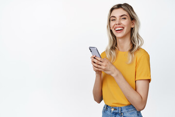 Naklejka premium Happy female model using mobile phone, smiling and laughing, standing with smartphone against white background