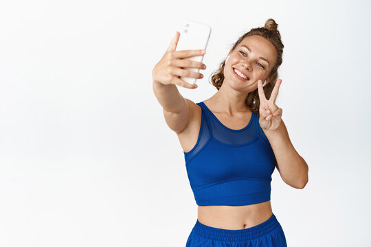 Happy female athlete taking selfie on mobile phone, photographing in headphones and activewear, white background