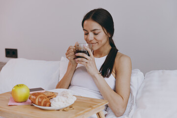 Amazing young woman in white tank top sit in bed have breakfast eat apple croissant hold in hand drink smell coffee in bedroom lounge home in own room house wake up dream be lost in reverie good day.