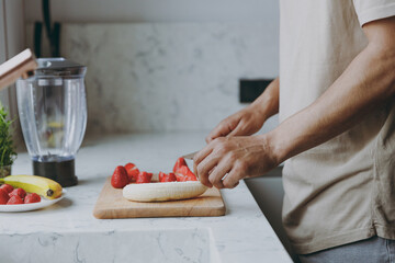 Cropped close up photo shot of young man 20s wear casual clothes look at window prepare sweet salad chop cut fruit cooking food in light indoor kitchen at home alone. Healthy diet lifestyle concept