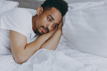Young pensive african american man in nightwear sleep with folded hands under cheek lying in bed rest relax spend time in bedroom lounge home in room house wake up dream be lost in reverie good day