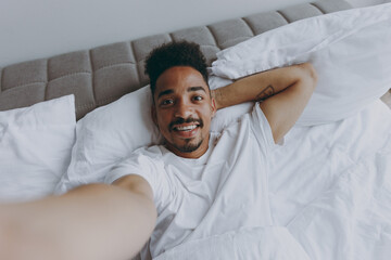 Close up young african american man in nightwear do selfie shot pov mobile phone lying in bed rest relax spend time in bedroom lounge home in own room house wake up dream be lost in reverie good day.