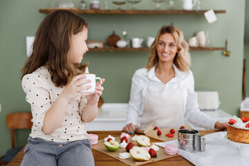 Happy fun chef cook baker mom woman wear white shirt work with smiling fun child baby girl helper drink tea at kitchen table home. Cooking food process concept Mommy little kid prepare fruit pie cake.