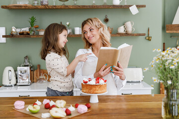 Lovely chef cook baker mom woman in white shirt work with baby girl helper read cookbook recipe at kitchen table home Cooking food process concept Mommy little kid daughter prepare fruit sweet cake.