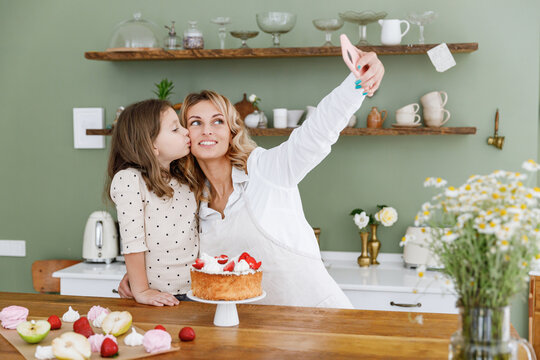 Happy chef cook baker mom woman in white shirt work baby girl helper do selfie shot on mobile cell phone kiss at kitchen table home Cooking food process concept Mommy little kid daughter prepare cake