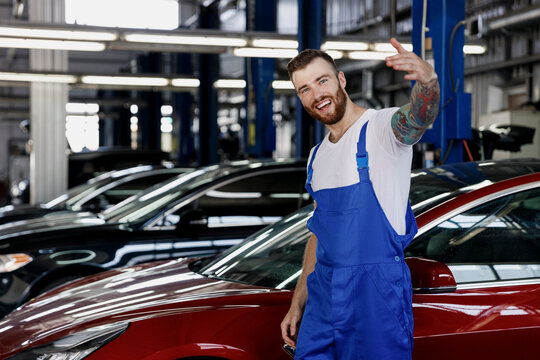 Strong smiling friendly happy young male professional technician car mechanic man wears denim blue overalls white t-shirt wave hand calling work in light modern vehicle repair shop workshop indoors.
