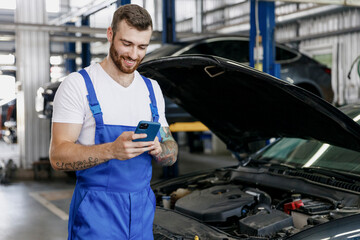 Young professional technician car mechanic man 20s in blue overalls white t-shirt use hold mobile...