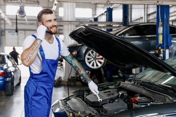 Young technician car mechanic man in denim overalls white t-shirt gloves listen music in earphones...