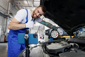 Fototapeta na wymiar Young troubleshooter technician car mechanic man in blue overalls white t-shirt talk mobile cell on phone stand show thumb up fix problem with raised hood work in vehicle repair shop workshop indoors