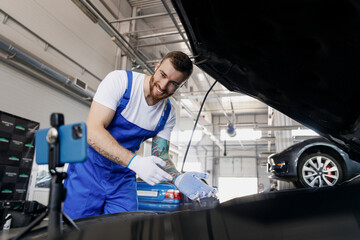 Young professional technician car mechanic man in blue overalls white t-shirt talk mobile cell on phone stand showing process fix problem with raised hood work in vehicle repair shop workshop indoors