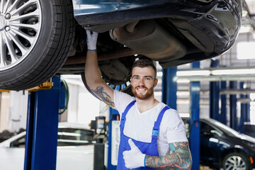 Strong happy young male professional technician mechanic man wears denim blue overalls t-shirt show thumb up stand near car lift check technical condition work in vehicle repair shop workshop indoors.