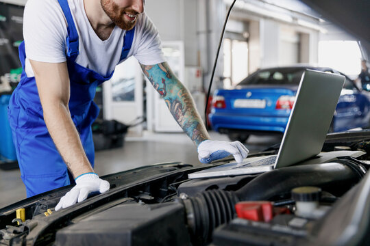 Cropped young professional technician car mechanic man in blue overalls t-shirt use laptop pc computer make diagnostics check fix problem with raised hood work in vehicle repair shop workshop indoor - Powered by Adobe