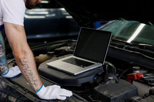 Repairman professional technician car mechanic man male hands use laptop pc computer make doing diagnostics check fix problem with raised hood bonnet working in vehicle repair shop workshop indoor