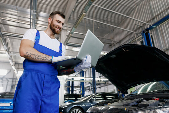 Lower young professional technician car mechanic man in blue overalls t-shirt use hold laptop pc computer make diagnostics fix problem with raised hood work in vehicle repair shop workshop indoor