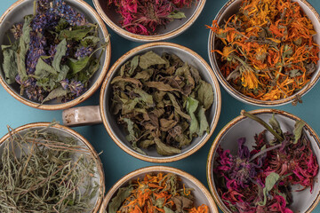Assortment of dried relaxing tea herbs in colourful cups on blue background close up. Calendula, mint, anise hyssop, monarda didyma, wormwood, sage leaves.