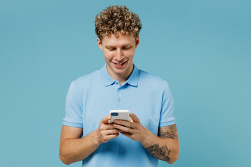 Happy smiling young curly man 20s years old wears azure t-shirt hold in hand use mobile cell phone chatting browsing send sms read news isolated on plain pastel light blue background studio portrait.