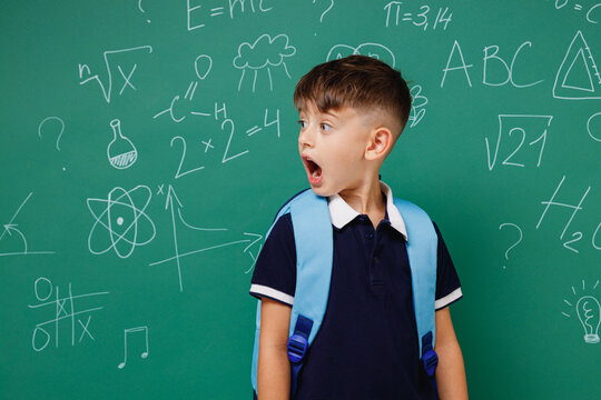 Young amazed surprised male kid school boy 5-6 years old in t-shirt backpack look aside isolated on green wall chalk blackboard background studio. Childhood children kids education lifestyle concept.