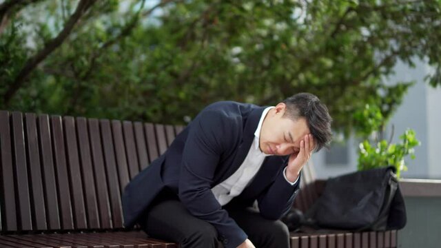 exhausted young asian business man office worker sitting on bench in city park outdoors. stressed overworked male with a headache massages head. Depressed Tired employe suffering pain sick work