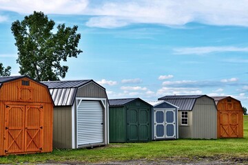 Various styles of wooden sheds on display. American shed is typically a simple, single-story roofed structure in a back garden or on an allotment that is used for storage, hobbies, or as a workshop. - obrazy, fototapety, plakaty